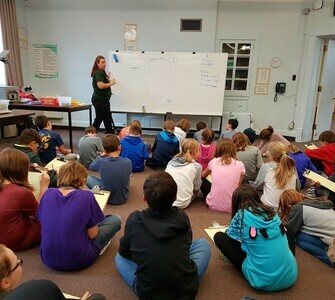 Students sitting on the f;oor looking at a whiteboard.